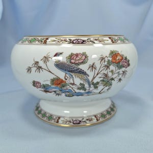 Puede incluir: Un cuenco de cer&aacute;mica blanca con base de pedestal, con un dise&ntilde;o detallado de p&aacute;jaros y flores en tonos azules, naranjas y rosas. El borde y la base est&aacute;n adornados con ribetes dorados y patrones decorativos. Una pieza decorativa para el hogar.