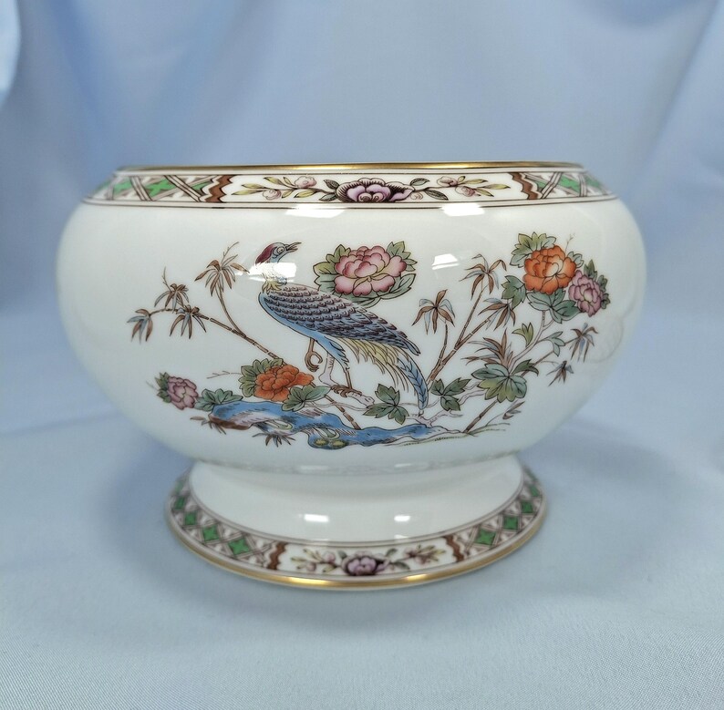 Puede incluir: Un cuenco de cer&aacute;mica blanca con base de pedestal, con un dise&ntilde;o detallado de p&aacute;jaros y flores en tonos azules, naranjas y rosas. El borde y la base est&aacute;n adornados con ribetes dorados y patrones decorativos. Una pieza decorativa para el hogar.