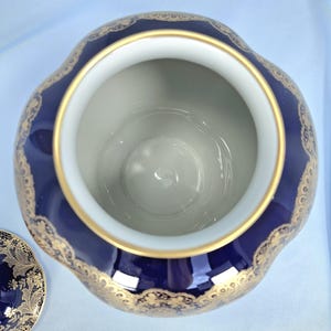 Może przedstawiać: Głęboko niebieski ceramiczny słoik z falistym brzegiem i złotą krawędzią wewnętrzną. Zewnętrzna strona ozdobiona jest misternym złotym wzorem filigranowym. Pasująca pokrywka z podobnym złotym wzorem koronki znajduje się obok słoika.