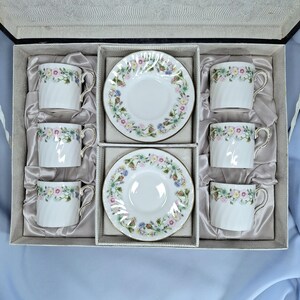 Peut inclure: Un coffret de six tasses et soucoupes en porcelaine blanche. Chaque tasse et soucoupe pr&eacute;sente un motif floral avec une bordure dor&eacute;e. L'ensemble est dispos&eacute; dans une bo&icirc;te grise avec une doublure en satin.