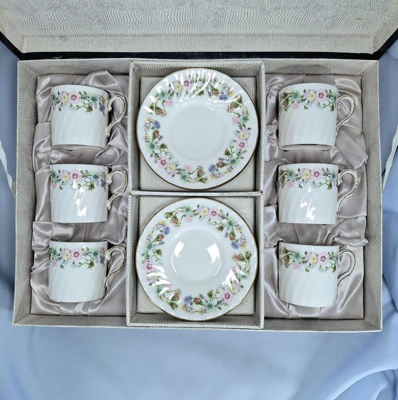 Peut inclure: Un coffret de six tasses et soucoupes en porcelaine blanche. Chaque tasse et soucoupe pr&eacute;sente un motif floral avec une bordure dor&eacute;e. L'ensemble est dispos&eacute; dans une bo&icirc;te grise avec une doublure en satin.
