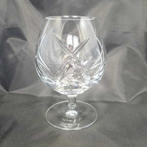 Puede incluir: Una copa de brandy de cristal transparente con un cuenco ancho y redondeado que presenta intrincados patrones de estrella tallados profundamente. El cristal tiene un tallo corto y robusto y una base circular con la marca John Rocha Waterford grabada.