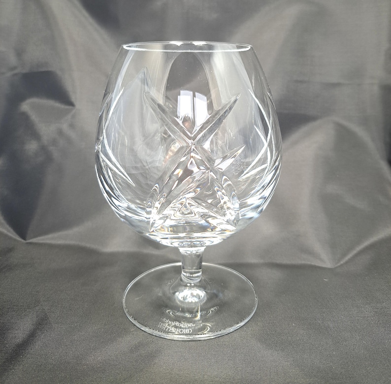 Puede incluir: Una copa de brandy de cristal transparente con un cuenco ancho y redondeado que presenta intrincados patrones de estrella tallados profundamente. El cristal tiene un tallo corto y robusto y una base circular con la marca John Rocha Waterford grabada.