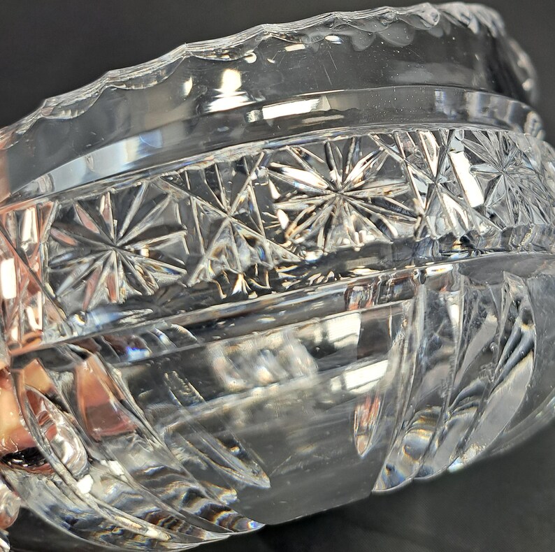 Op de afbeelding: Een heldere glazen kom met een decoratief geslepen patroon met sterrenmotieven rondom het bovenste gedeelte en verticale groeven die naar de basis toe lopen. De rand heeft een geschulpte afwerking en het glas reflecteert het licht.