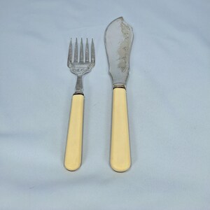 May include: A vintage fork and butter knife set. The fork has four tines and a cream-colored handle. The butter knife has a curved blade with decorative etching and a matching cream-colored handle. The set is made of silver-colored metal.
