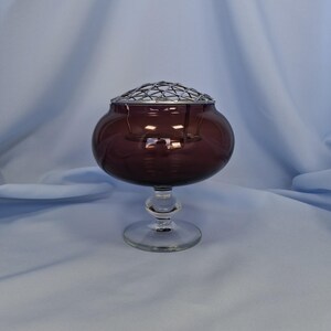 Puede incluir: Un jarr&oacute;n de flores de cristal morado oscuro con base y tallo de cristal transparente. El jarr&oacute;n tiene forma de cuenco redondo y una rejilla met&aacute;lica en la parte superior. El jarr&oacute;n est&aacute; dise&ntilde;ado para contener flores y es un objeto decorativo.