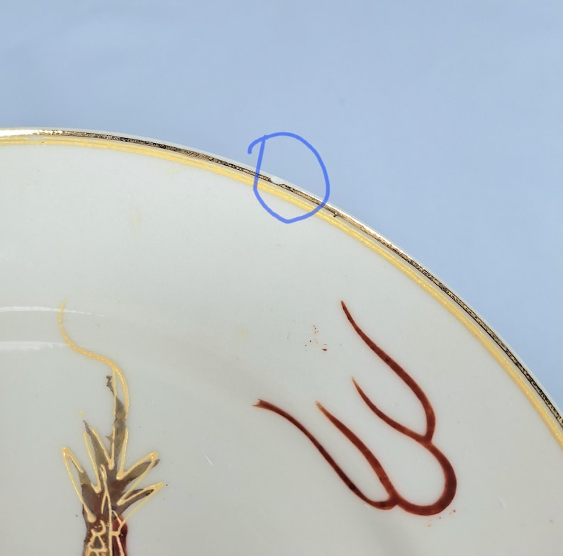 May include: Close-up of a white ceramic plate with a gold rim and decorative brown and gold accents. The plate features a stylized design, possibly floral or abstract, with a gold border. The plate is set against a light blue background.