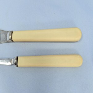 May include: Two antique butter knives with ivory-colored handles and silver blades. The handles have a smooth, rounded shape. The knives are set against a light blue background.