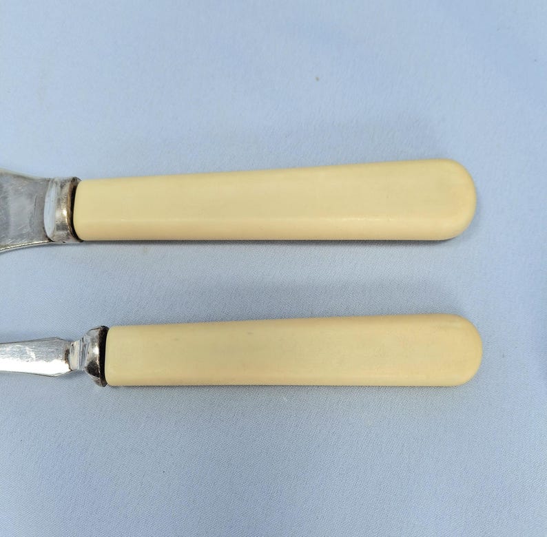 May include: Two antique butter knives with ivory-colored handles and silver blades. The handles have a smooth, rounded shape. The knives are set against a light blue background.