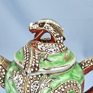 Pu&ograve; includere: Primo piano di una teiera in ceramica con un motivo a drago. La teiera presenta un corpo verde con dettagli bianchi e marroni, una testa di drago rossa sul coperchio e finiture dorate. Anche il manico e il beccuccio sono decorati con motivi intricati.