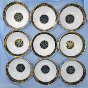 Op de afbeelding: Een set van negen witte porseleinen kommen gerangschikt in een drie-bij-drie raster. Elke kom heeft een brede donkerblauwe rand met gouden filigraanpatronen en een bijpassend rond donkerblauw en goud bloemenmedaillon op de bodem.