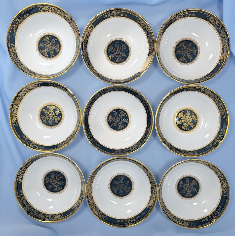 Op de afbeelding: Een set van negen witte porseleinen kommen gerangschikt in een drie-bij-drie raster. Elke kom heeft een brede donkerblauwe rand met gouden filigraanpatronen en een bijpassend rond donkerblauw en goud bloemenmedaillon op de bodem.