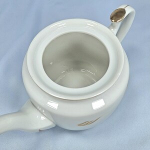 May include: Overhead view of a white ceramic teapot with a gold accent on the handle and a gold leaf design. The teapot has a round body and a spout extending from the side. The lid is open, revealing the interior.