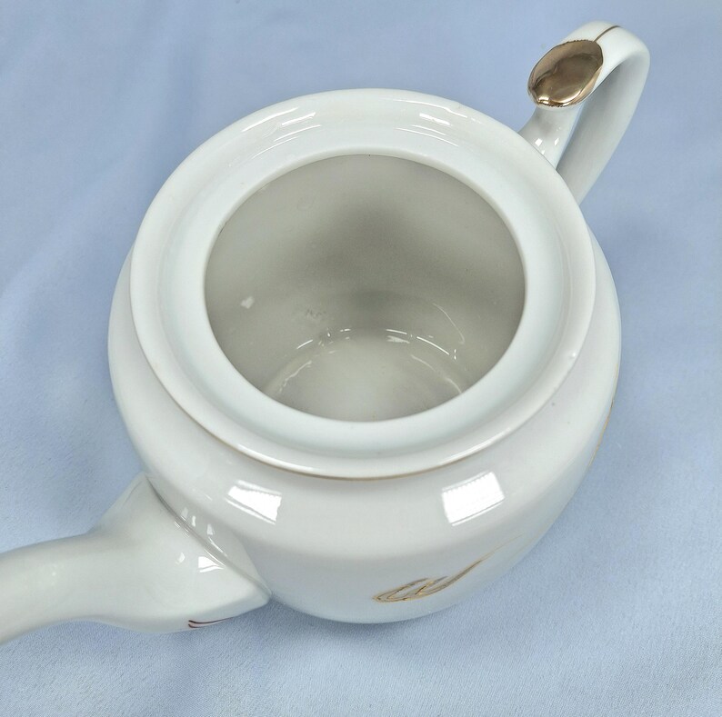 May include: Overhead view of a white ceramic teapot with a gold accent on the handle and a gold leaf design. The teapot has a round body and a spout extending from the side. The lid is open, revealing the interior.