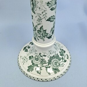 K&ouml;nnte beinhalten: Wei&szlig;e Keramikvase mit ausgestelltem Fu&szlig; und langem, schlankem Hals. Die Vase ist mit einem gr&uuml;nen Blumenmuster verziert, das Bl&auml;tter und Bl&uuml;ten umfasst. Der Sockel hat einen dekorativen Rand.