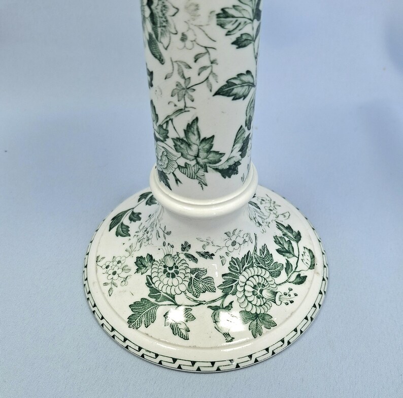K&ouml;nnte beinhalten: Wei&szlig;e Keramikvase mit ausgestelltem Fu&szlig; und langem, schlankem Hals. Die Vase ist mit einem gr&uuml;nen Blumenmuster verziert, das Bl&auml;tter und Bl&uuml;ten umfasst. Der Sockel hat einen dekorativen Rand.
