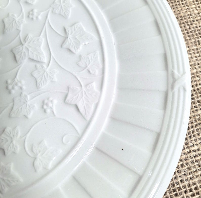 K&ouml;nnte beinhalten: Wei&szlig;e Keramikplatte mit gepr&auml;gtem Efeublatt- und Beerendesign. Die Platte hat ein detailliertes Reliefmuster aus Bl&auml;ttern und Beeren, mit einem gerippten Rand und dekorativen Akzenten. Ein klassisches und elegantes St&uuml;ck zum Servieren oder Ausstellen.