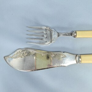 May include: Silverware set featuring a fork and a butter knife. The fork has four tines and a decorative handle. The butter knife has a curved blade and a matching handle. Both pieces have off-white handles and ornate detailing.