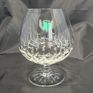 May include: A clear crystal brandy snifter featuring a wide, rounded bowl with a deep diamond-cut pattern and a short, thick stem. The circular base displays a radiating starburst design. A small, round green sticker is attached to the glass.