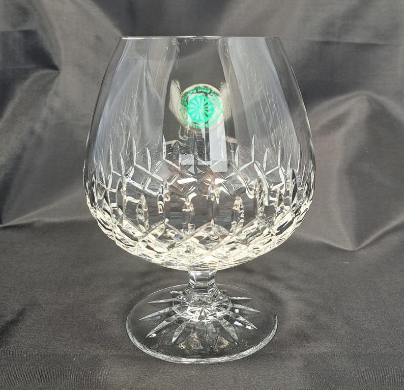 May include: A clear crystal brandy snifter featuring a wide, rounded bowl with a deep diamond-cut pattern and a short, thick stem. The circular base displays a radiating starburst design. A small, round green sticker is attached to the glass.