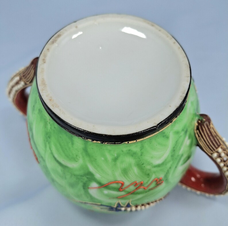 Pu&ograve; includere: Una zuccheriera in ceramica verde con interno bianco, bordo nero e due manici. La ciotola presenta un disegno dipinto con accenti rossi e blu. I manici sono marroni con dettagli bianchi a forma di perla e accenti rossi.