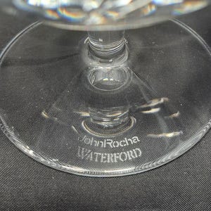 Puede incluir: Una base de cristal transparente de una pieza de cristaler&iacute;a con un pie circular. El cristal tiene grabado el texto John Rocha y WATERFORD en la superficie. El tallo se eleva desde el centro de la base circular, de acabado liso y transparente.