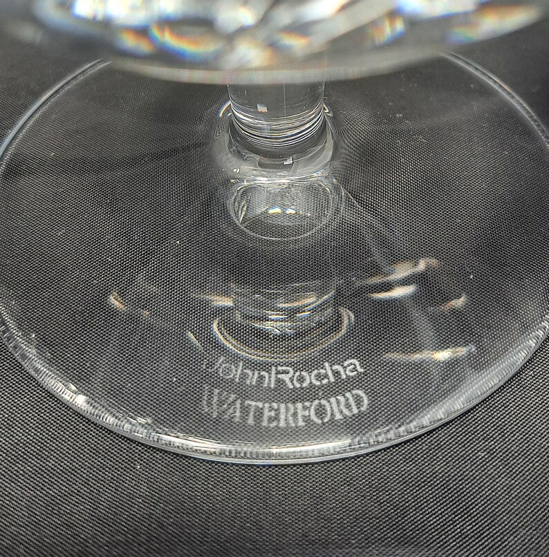 Puede incluir: Una base de cristal transparente de una pieza de cristaler&iacute;a con un pie circular. El cristal tiene grabado el texto John Rocha y WATERFORD en la superficie. El tallo se eleva desde el centro de la base circular, de acabado liso y transparente.