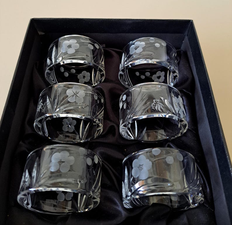 May include: A set of six clear glass napkin rings arranged in a black box. Each circular ring features a delicate etched floral pattern with five-petaled blossoms and slender leaves, creating a translucent design on the smooth glass surface.