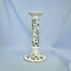 K&ouml;nnte beinhalten: Ein wei&szlig;er Kerzenhalter aus Keramik mit einem gr&uuml;nen Blumenmuster. Der Kerzenhalter hat einen breiten Sockel, einen hohen, zylindrischen Schaft und eine kleine, kreisf&ouml;rmige Oberseite. Das Blumenmuster umfasst Bl&auml;tter und Bl&uuml;ten.