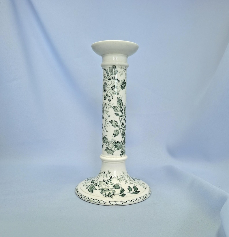K&ouml;nnte beinhalten: Ein wei&szlig;er Kerzenhalter aus Keramik mit einem gr&uuml;nen Blumenmuster. Der Kerzenhalter hat einen breiten Sockel, einen hohen, zylindrischen Schaft und eine kleine, kreisf&ouml;rmige Oberseite. Das Blumenmuster umfasst Bl&auml;tter und Bl&uuml;ten.