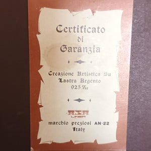 Puede incluir: Un certificado de garant&iacute;a para una creaci&oacute;n de plata. El certificado est&aacute; impreso en papel crema con bordes marrones. El texto dice "Certificato di Garanzia", "Creazione Artistica Su Lastra Argento 925%", "LINEA", "marchio preziosi AN-22" y "Italy".