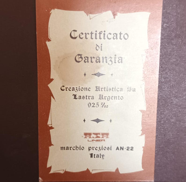 Puede incluir: Un certificado de garant&iacute;a para una creaci&oacute;n de plata. El certificado est&aacute; impreso en papel crema con bordes marrones. El texto dice "Certificato di Garanzia", "Creazione Artistica Su Lastra Argento 925%", "LINEA", "marchio preziosi AN-22" y "Italy".