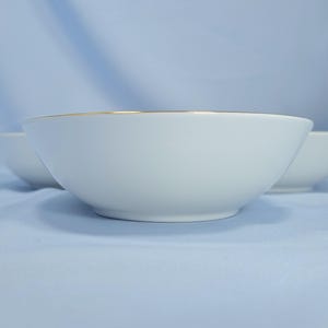 Op de afbeelding: Een witte keramische kom met een gladde, ronde vorm en een dunne gouden rand langs de bovenkant. De kom heeft een strak ontwerp met een effen witte afwerking en een metalen gouden accent dat de gehele bovenste omtrek van de kom omcirkelt.
