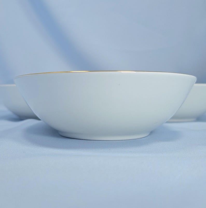 Op de afbeelding: Een witte keramische kom met een gladde, ronde vorm en een dunne gouden rand langs de bovenkant. De kom heeft een strak ontwerp met een effen witte afwerking en een metalen gouden accent dat de gehele bovenste omtrek van de kom omcirkelt.