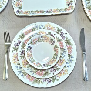 以下が含まれることがあります： 白地にピンク、黄色、紫の花柄が施されたフォーマルな食器セット。セットには、お皿、サービングトレイ、フォーク、ナイフが含まれています。お皿とトレイの縁は金色のトリムです。