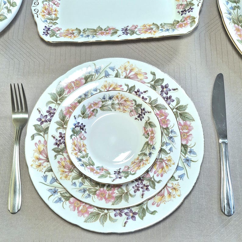 以下が含まれることがあります： 白地にピンク、黄色、紫の花柄が施されたフォーマルな食器セット。セットには、お皿、サービングトレイ、フォーク、ナイフが含まれています。お皿とトレイの縁は金色のトリムです。