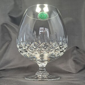 May include: A clear crystal brandy snifter featuring a wide, rounded bowl with a diamond-cut pattern on the lower half and a short, faceted stem leading to a circular base. A small green circular sticker is attached to the upper rim.