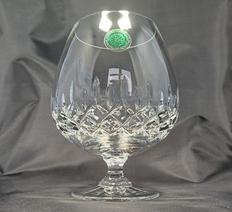 May include: A clear crystal brandy snifter featuring a wide, rounded bowl with a diamond-cut pattern on the lower half and a short, faceted stem leading to a circular base. A small green circular sticker is attached to the upper rim.