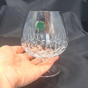May include: A clear crystal brandy snifter glass featuring a wide, rounded bowl with a deep diamond-cut pattern on the lower half. A small circular green sticker with white text is adhered to the upper side of the glass, which has a short stem.