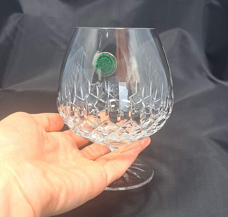 May include: A clear crystal brandy snifter glass featuring a wide, rounded bowl with a deep diamond-cut pattern on the lower half. A small circular green sticker with white text is adhered to the upper side of the glass, which has a short stem.
