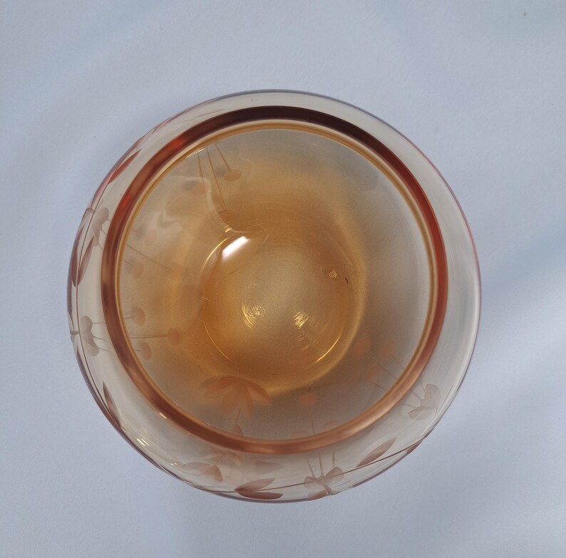 May include: A top-down view of a round, amber-colored glass bowl featuring a delicate, etched floral pattern around its exterior rim. The glass has a smooth, translucent finish, and the circular opening reveals the deep, warm orange interior.