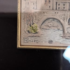 Puede incluir: Una obra de arte enmarcada en plata que representa un puente sobre un r&iacute;o con edificios en el fondo. El marco tiene una placa de plata con el texto "R 1+2g. 925" y el a&ntilde;o "1722 AN".