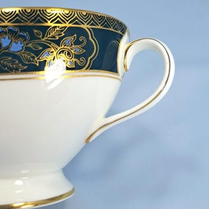 May include: A white porcelain teacup featuring a dark teal band around the upper rim, decorated with gold filigree patterns and light blue floral motifs. The cup includes a curved handle with gold trim and a gold-lined base.