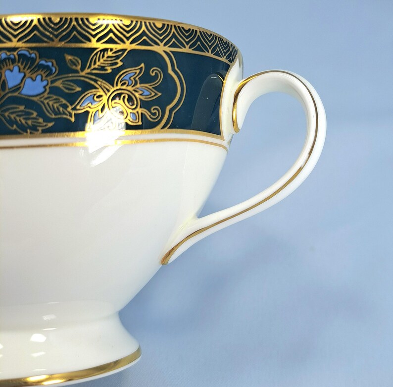 May include: A white porcelain teacup featuring a dark teal band around the upper rim, decorated with gold filigree patterns and light blue floral motifs. The cup includes a curved handle with gold trim and a gold-lined base.