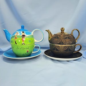 May include: Two tea-for-one sets. The left set is light blue and green with illustrations of two figures in kilts and the text 'Bonnie Scotland...'. The right set is black with a gold fan-shaped pattern and a metallic gold lid and handle.