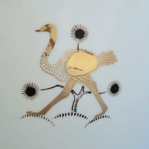 May include: A decorative illustration of an ostrich in gold and brown tones, set against a white background. The ostrich is depicted with detailed gold accents, surrounded by black sunburst-like shapes and a stylized tree.