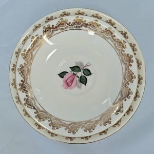 May include: A set of three white porcelain plates. The plates have a gold filigree border and a pink rose with green leaves in the centre. The plates are stacked on top of each other.
