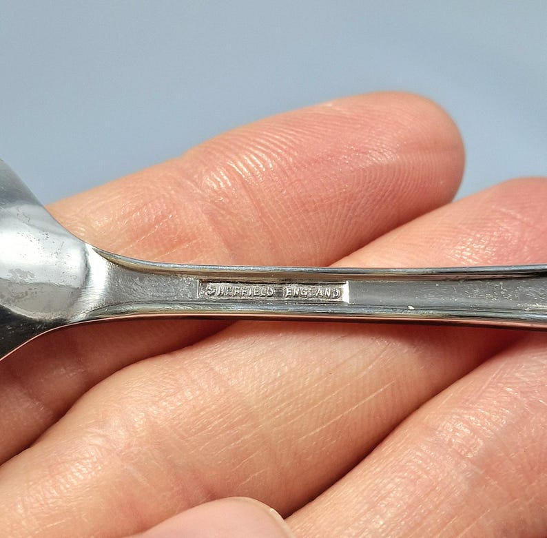 May include: Close-up of a silver spoon with a rounded bowl. The spoon's handle has the text "Sheffield England" engraved on it. The spoon is held in a person's hand.