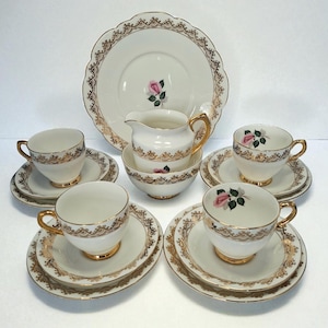 May include: A vintage tea set with a cream-colored base and gold trim. The set includes teacups, saucers, a creamer, a sugar bowl, and a large serving plate. Each piece features a pink rose design, perfect for a tea party.