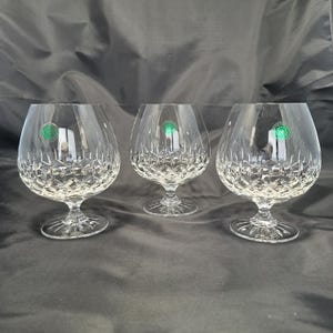 May include: Three clear crystal brandy glasses featuring a deep diamond-cut pattern on the lower bowls and short, sturdy stems with a starburst design on the circular bases. Each glass displays a small, round green sticker on the upper side.